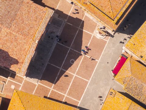 Aerial view of terracotta rooftops casting stark shadows on the Piazza Pio II, where people gather near the well, creating a vibrant contrast of light and shade, Pienza, Tuscany, Italy.