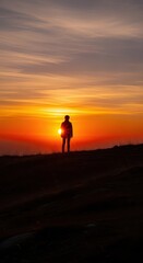 Fototapeta premium Solitary figure stands silhouetted against brilliant orange sunset atop a darkened hillside