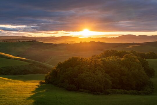 Aerial view of the sun kissing the rolling green hills and distant farmhouses under a dramatic sky, Crete Senesi, Italy.