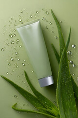 Green tube of skincare product placed on green background with fresh aloe vera leaves and water droplets