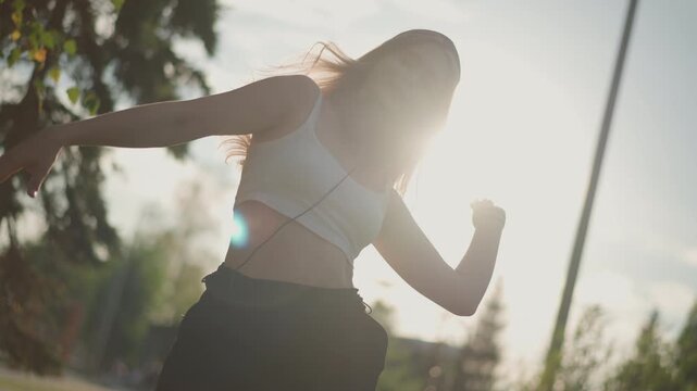 Caucasian woman dancing street golden hour, backlit silhouette with headphones and beanie, crop top and relaxed pants, energetic arm movements and head bops, sunflare through leaves, leafy urban park