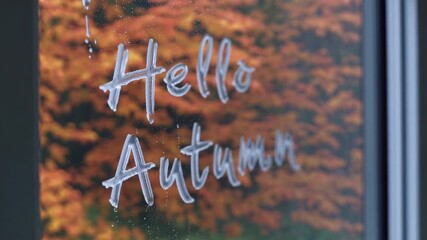 The phrase hello autumn is written on a rain-speckled window, with a backdrop of vibrant fall foliage. the image portrays a warm, welcoming ambiance complemented by the rich colors of autumn leaves, p