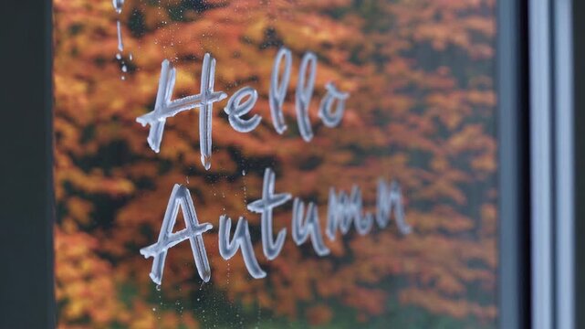 The phrase hello autumn is written on a rain-speckled window, with a backdrop of vibrant fall foliage. the image portrays a warm, welcoming ambiance complemented by the rich colors of autumn leaves, p