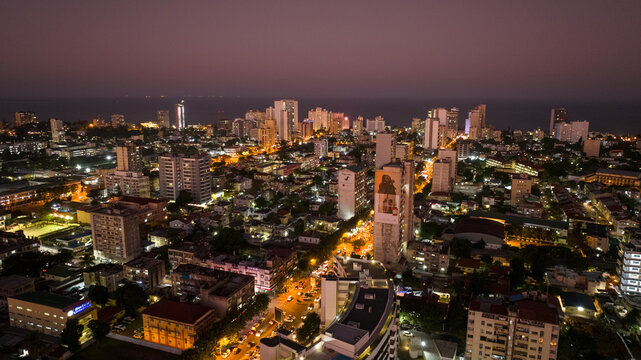 Aerial view of the illuminated cityscape meeting the dark ocean horizon under a purple sky, showcasing the contrast of light and shadow, Maputo, Cidade de Maputo, Mozambique.