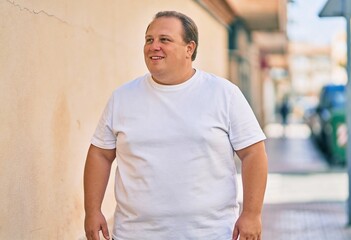 A person is standing outdoors wearing a plain white t-shirt, looking to the side with a smile, in a brightly lit urban setting with buildings and a sidewalk in the background