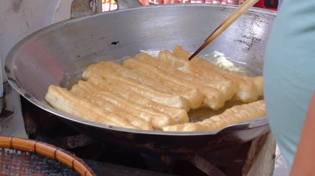 Close-up of a street food vendor frying Youtiao or Yu Char Kway (Cakwe) in the hot oily pan. it is a long golden-brown deep-fried strip of wheat flour dough of Chinese origin.