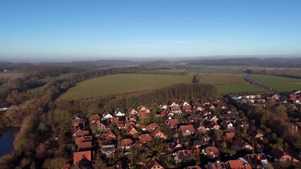 Aerial flyover shot of German suburban neighborhood with red-roofed houses beside open fields, on clear winter day with light frost and low sun over the rural landscape. Wide shot. Fog in distance.