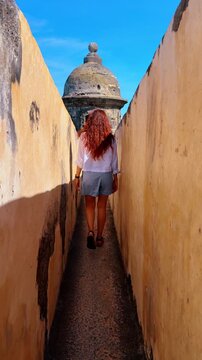 Woman Walking in Narrow Fortress Passage Towards Sentry Box. Female tourist walks through a narrow stone corridor inside a historic fortress leading towards a domed sentry box against a blue sky.