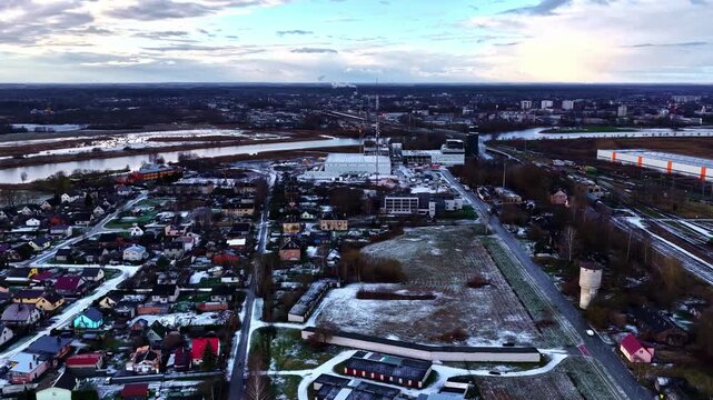 Drone aerial over winter suburbs beside a river and industrial complex, with patchy snow fields, highway corridor, and a tall broadcast tower under moody clouds.
