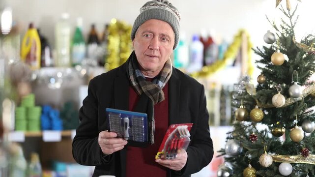Mature man pulls a razor and a hair trimmer in his hands and chooses which one to take. Man buying razors on the background of a Christmas tree in a supermarket. High quality 4k footage