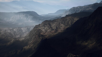 Layers of rugged mountains stretch across the landscape, shrouded in a gentle mist. The soft light highlights the textures and shapes of the terrain, creating an ethereal atmosphere.