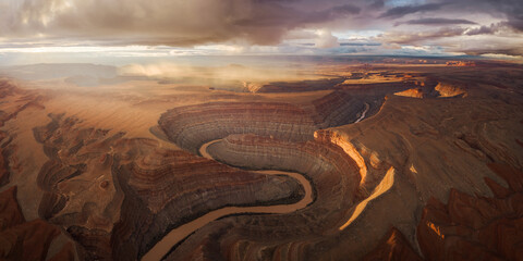 Aerial view of a dramatic river carves its way through the rugged, sun-kissed landscape under a sky alive with stormy hues, Goosenecks State Park, Bluff, Utah, United States.