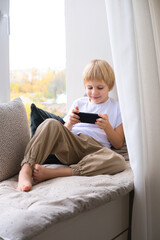 blonde little boy using smartphone sitting on the windowsill