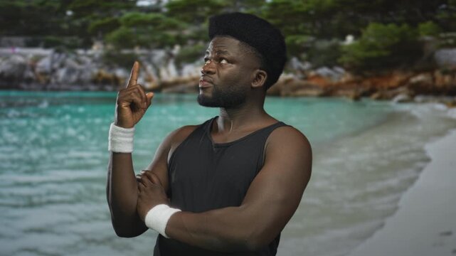 Man with bare arms and white wristbands points index finger upward on beach with crossed arm pose and focused expression; confidence determination.