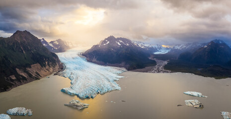 Aerial view of the majestic blue glacier flowing into a calm lake dotted with icebergs, cradled by rugged mountains under a dramatic sky, Cordova, Alaska, United States.