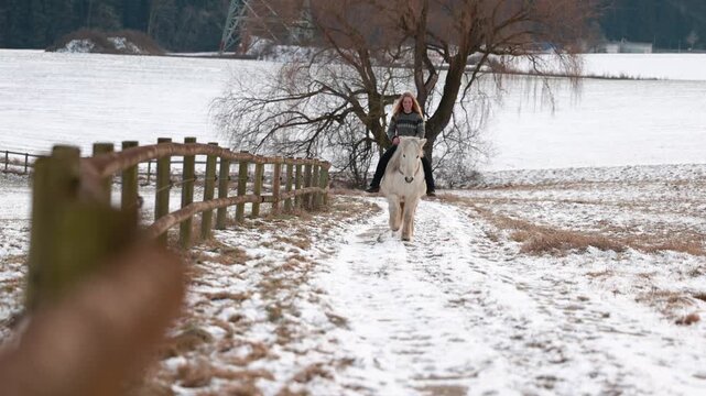 A young rider rides her white Icelandic gelding with a neck ring on a snowy rural path, calm winter scene outdoors