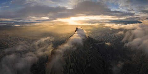 Aerial view of a rugged, dark mountain peak piercing through a sea of ethereal clouds, bathed in the golden light of the setting sun, Hanksville, Utah, United States.
