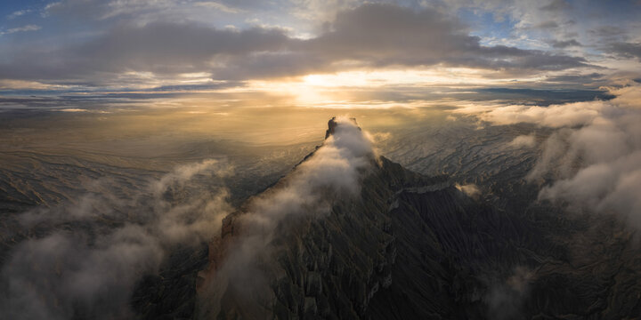 Aerial view of a rugged, dark mountain peak piercing through a sea of ethereal clouds, bathed in the golden light of the setting sun, Hanksville, Utah, United States.