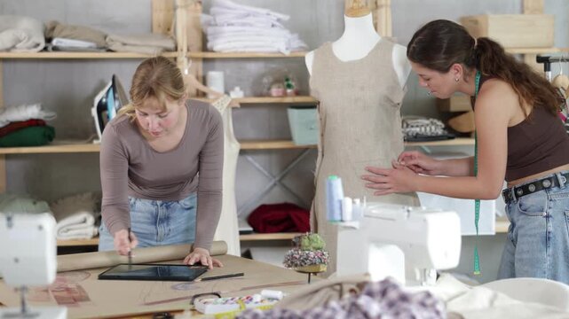 Thoughtful young female dressmaker working on digital sketch of new dress using tablet in fashion studio while colleague working on fitting dress on mannequin in background