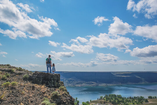 Rear view of a father standing on a mountain ledge holding his daughters hand and looking down at a lake in summer