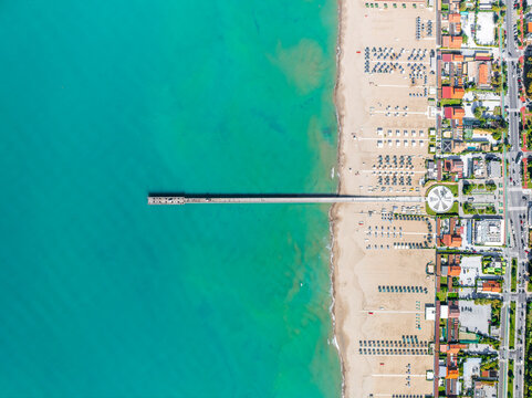 Aerial view of a pier stretching into the turquoise sea, beach umbrellas dotting the golden sand meeting the neat urban grid, Forte dei Marmi, Tuscany, Italy.