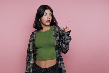 Young woman in a green top and plaid shirt points in a bright pink room while making an expressive...