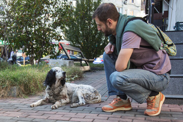 Adult man crouching on a city sidewalk watching a homeless dog resting on the pavement, calm moment of empathy and compassion, urban street scene about kindness, care and animal welfare