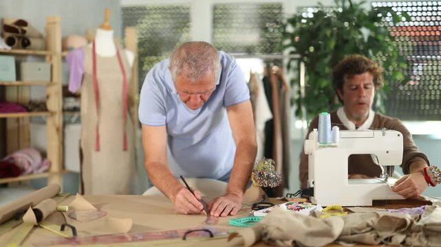 Adult man tailor sews on machine elderly man assistant drawing pattern on paper in workshop