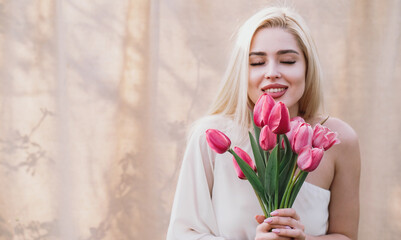 A blonde girl with long hair holding a bouquet of pink tulips against a light textured background. Happy International Women's Day, the first day of spring, Mother's Day, and spring holiday.