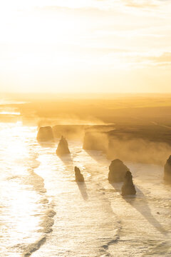 Aerial view of the Twelve Apostles rock formations shrouded in a golden haze, waves crashing gently against the shore, Lorne, Victoria, Australia.