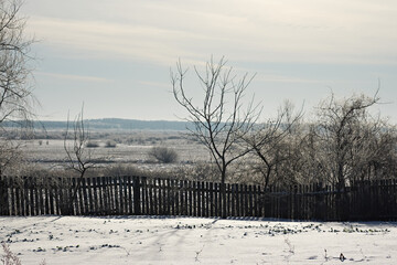 The image shows a rural winter landscape. A wooden fence separates the foreground from a snowy field in the background. The leafless trees are covered in a light frost.