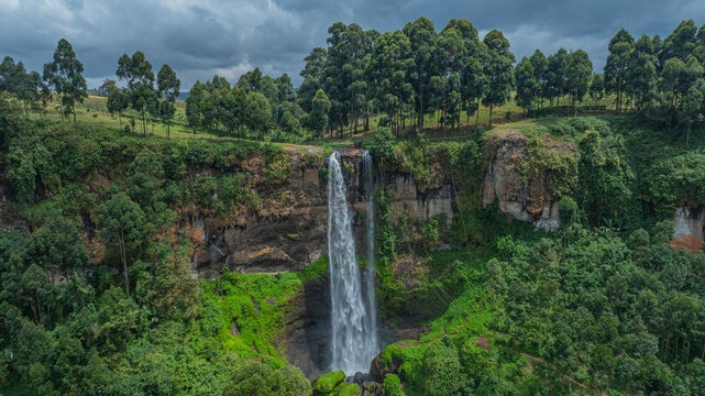 Aerial view of a majestic waterfall cascading down a rocky cliff face, surrounded by lush green vegetation under a cloudy sky, Sipi, Eastern Region, Uganda.