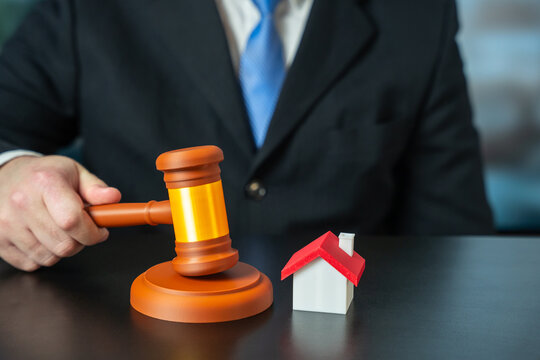 Male hand holds a judge&rsquo;s gavel near house. Symbolizing real estate legal decisions, property inheritance disputes, and the establishment of ownership rights.