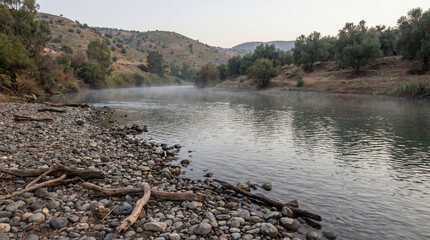 Misty river landscape at dawn with rocky bank and driftwood. Scenic water flow through hills resembling the biblical Jordan River