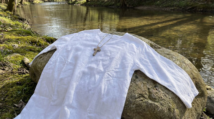 White linen robe and wooden cross on a rock by the river. Christian baptism of Jesus concept. Religious symbol of faith and purification
