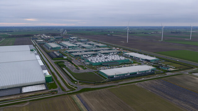 Aerial view of sprawling industrial buildings punctuated by rows of cylindrical structures, with wind turbines standing tall in the distance, Middenmeer, Netherlands.