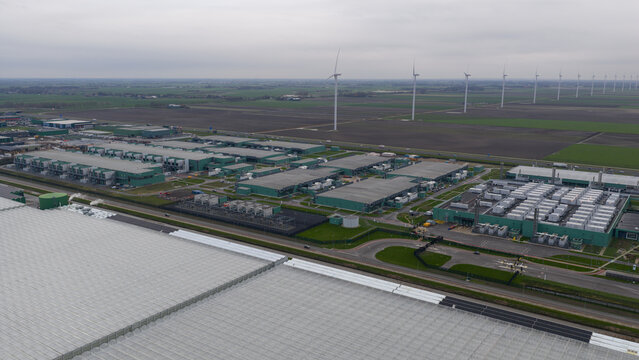 Aerial view of a vast data center complex juxtaposed against expansive greenhouses and distant wind turbines under a muted sky, AI Data Center, Middenmeer, Netherlands.