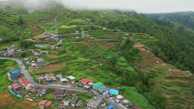 High forward aerial gliding above the winding mountain road, revealing dense rice terraces and colorful settlements in Buguias, Benguet, Philippines.