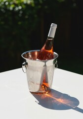 Refreshing chilled rose wine bottle nestled in an ice bucket, casting dramatic shadows on a clean white outdoor table in bright sunlight, sophisticated, white, table