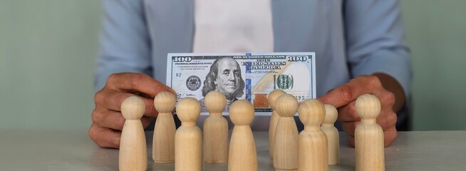 Woman holds a hundred dollar bill in front of wooden figures representing different groups of...