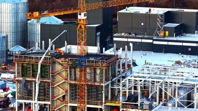 Drone close-up of crane and workers on snowy industrial construction site with concrete columns, steel framework, and scaffolding during winter building project.