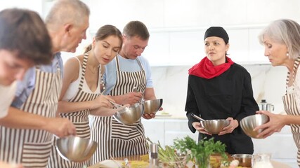 Positive young Asian woman professional chef conducting culinary courses, standing next to table with carafe of cream, imparting cooking skills to diverse group of people of different ages. High