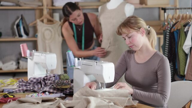 Female tailor is working on a sewing machine, checking the neatness of the stitches against the background of an assistant and a mannequin. Two tailors in a workshop