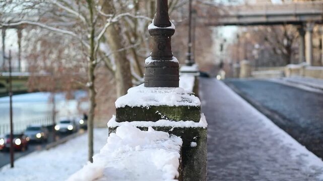 Winter scene in Paris with a close-up of a traditional lamp post covered in snow along a cobblestone street. A car with headlights on is driving in the background