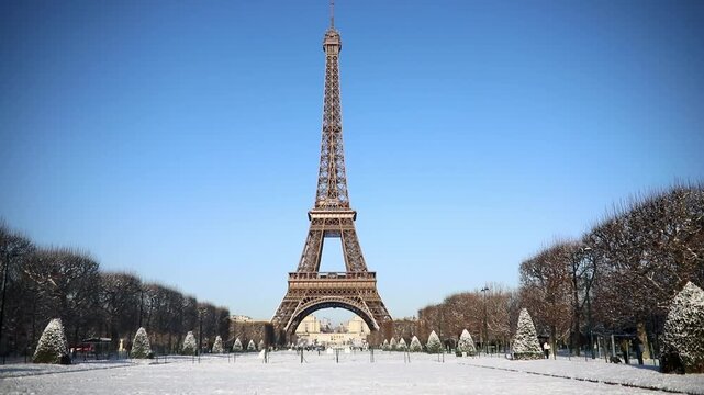 Front view of the Eiffel Tower rising above a snow-covered park in Paris on a sunny winter day. Blue sky, snowy grounds, and trimmed trees create a postcard-perfect scene.