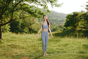 Young woman practicing yoga in nature, wearing stylish gray activewear, exuding calmness and serenity amidst a lush green background, perfect for wellness themes