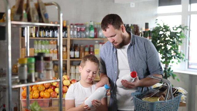  Man with son hold packages bottles and choose consider milk in store. Market visitor choose consider make choice bottle milk. Client hesitates before buying, chooses drink, reads labels on bottles