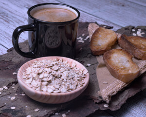 Black mug with coffee foam on tree bark, with a bowl of rolled oats and crispy toasted bread, rustic style