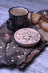 Black mug with coffee foam on tree bark, with a bowl of rolled oats and crispy toasted bread, rustic style