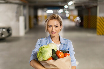 Fototapeta premium A woman smiles while holding a bag of fresh produce in a parking garage, ready to head home